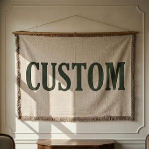 以下が含まれることがあります： ベージュ色の織り壁掛けで、ダークグリーンのブロック文字で「CUSTOM」と書かれています。 バナーは木の棒から吊り下げられており、縁はフリンジになっています。 バナーの背後にある壁は白です。