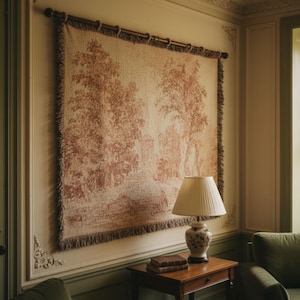 May include: A large tapestry depicting a pastoral scene in shades of cream and brown, mounted on a wooden rod. The tapestry is fringed along the bottom edge and hangs above a small wooden table with a lamp and books.