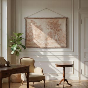 May include: A tapestry depicting a pastoral scene in shades of beige and brown hangs on a white wall. The tapestry is suspended by a wooden rod and a string. The room also features a wooden desk, a chair, and a small round table.