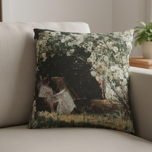 Pode incluir: Almofada decorativa quadrada com uma imagem impressa de uma pintura que retrata uma cena sob uma &aacute;rvore com flores brancas. A almofada tem um fundo escuro com figuras em roupas brancas. A almofada est&aacute; em um sof&aacute; bege.
