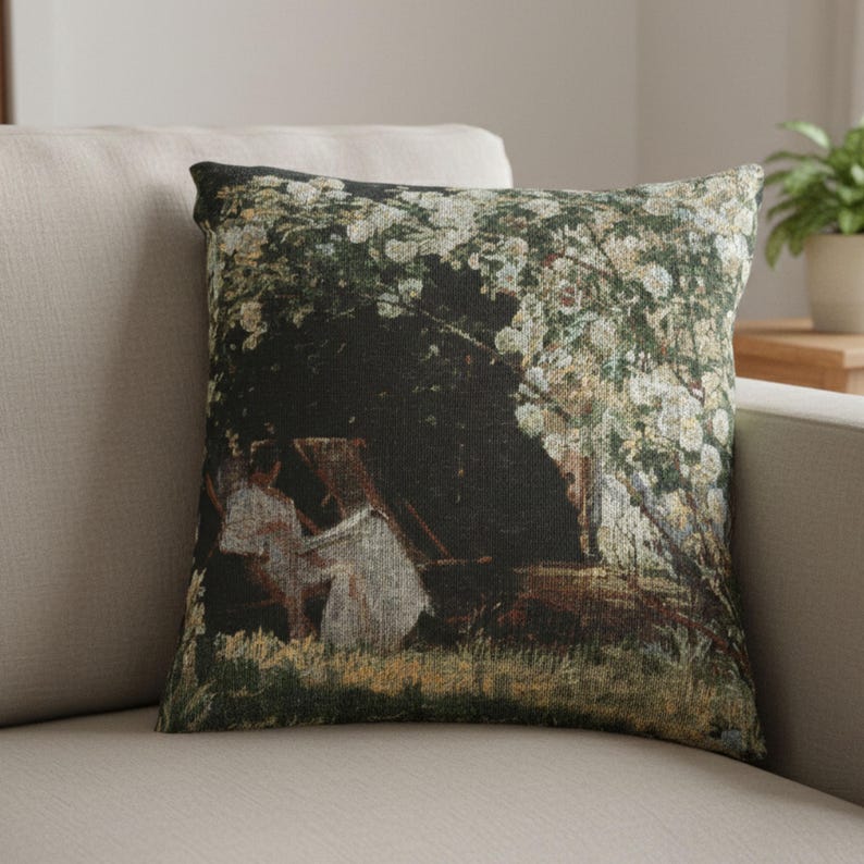 Pode incluir: Almofada decorativa quadrada com uma imagem impressa de uma pintura que retrata uma cena sob uma &aacute;rvore com flores brancas. A almofada tem um fundo escuro com figuras em roupas brancas. A almofada est&aacute; em um sof&aacute; bege.