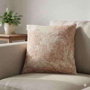 May include: Square decorative pillow with a textured, abstract design in shades of peach and cream. The pillow is placed on a neutral-colored sofa, creating a cozy and inviting atmosphere. The pillow's pattern resembles a watercolor painting.