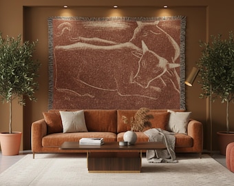 May include: A woven tapestry featuring a brown and cream-colored illustration of two cows. The tapestry is displayed above a brown sofa with cream-colored pillows. The room has a modern, minimalist design with olive trees in terracotta pots.