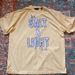 May include: A light yellow t-shirt with the words "be SALT & LIGHT" in blue and white stitched lettering. The shirt has a crew neck and short sleeves. The date "5 13 16" is below the word "LIGHT".