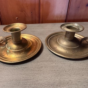 May include: Two antique brass candle holders. Each has a circular base, a raised candle cup, and a small handle. The brass has a warm, aged patina, suggesting they are vintage pieces. The candle holders are likely from the Victorian era.
