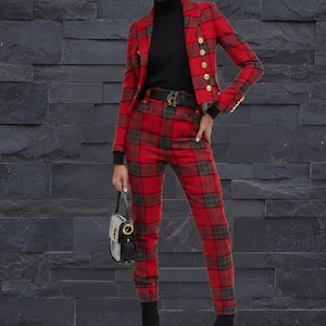 Puede incluir: Un conjunto de blazer y pantalón a cuadros rojos con botones dorados. El atuendo incluye un cuello alto negro, un cinturón negro con hebilla dorada y un pequeño bolso de mano en blanco y negro. El estampado a cuadros incluye acentos verdes.