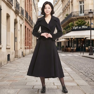 May include: A woman wearing a long, black coat with a V-neckline and a flared skirt, standing on a cobblestone street. The coat has a double-breasted design and is paired with black shoes. The background features a European street scene.