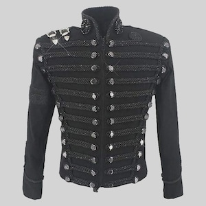 Op de afbeelding: Zwarte jas in militaire stijl met een hoge kraag en decoratieve touwversiering. De jas heeft meerdere rijen zilverkleurige knopen en versieringen op de schouders en kraag. De jas met lange mouwen heeft een getailleerde pasvorm.