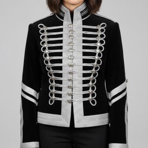 Könnte beinhalten: Schwarze Samtjacke mit silberner Verzierung und dekorativen Knöpfen. Die Jacke hat einen hohen Kragen mit gestickten Details und silbernen Akzenten an der Vorderseite und den Ärmeln. Ein stilvolles Kleidungsstück im Militärstil.