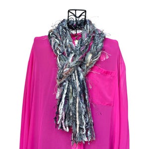 May include: A hand-woven scarf with a braided pattern, incorporating grey, white, and black threads with fringing. The scarf is shown on a black mannequin head over a vibrant pink blouse.