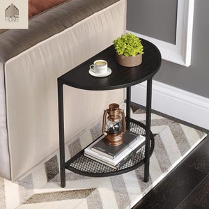 May include: Black, half-moon side table with a metal frame and two shelves. The top shelf holds a small potted plant and a cup of coffee. The lower shelf has a metal mesh design and holds books and a lantern. The table is placed on a patterned rug.