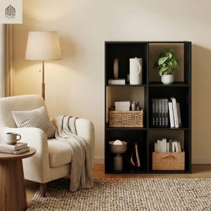 May include: A black cube storage unit with six compartments, each holding various decorative items. A white armchair with a patterned pillow and a beige throw blanket sits to the left. A floor lamp with a beige lampshade is next to the armchair.