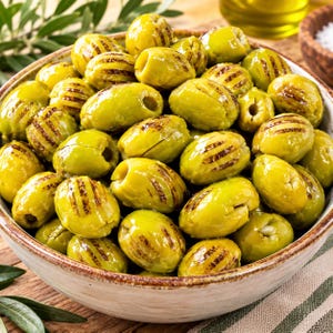 May include: A ceramic bowl filled with numerous pitted green olives, each featuring distinct dark brown char marks from grilling. The olives are oval-shaped, glossy, and piled high in the bowl, showcasing a vibrant light green color and texture.