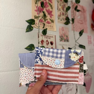 May include: A patchwork wallet with a variety of fabric patterns, including floral, gingham, and striped designs. The wallet has a light blue button closure and a small key ring attachment. The wallet is held in a hand.