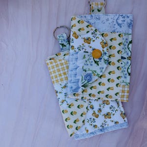 May include: Two fabric keychains with floral and checkered patterns. One keychain has a flap with a yellow button closure. The fabric features yellow, blue, and white floral designs. The keychains are on a pink background.