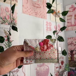 May include: A small, patchwork fabric wallet with a floral pattern in shades of red, pink, and beige. The wallet has a flap closure secured by a pink button. The design includes a variety of fabric patterns and textures.