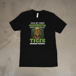 May include: Black t-shirt with a tiger graphic and the text "DAS IST MEIN MENSCHENKOSTÜM EIGENTLICH BIN ICH EIN TIGER NAMENS MARCEL". The tiger graphic is orange and brown, surrounded by green foliage.