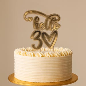 Peut inclure: Un gâteau rond avec un glaçage blanc et un décor de gâteau doré qui dit "hello 30" avec un cœur. Le gâteau est sur un présentoir à gâteau doré.