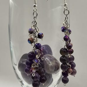 May include: Dangle earrings featuring clusters of purple and dark-coloured beads. The earrings hang from silver-coloured hooks and chains. The beads vary in size and shade, resembling grapes. The earrings are displayed in a clear glass.