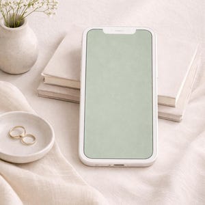 Puede incluir: Un teléfono inteligente blanco con una pantalla verde claro descansa sobre una tela color crema. Dos anillos dorados se encuentran en un plato blanco pequeño y redondo. Un pequeño jarrón blanco contiene delicadas flores blancas, y dos libros blancos están en el fondo.