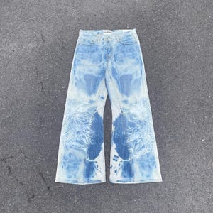 Op de afbeelding: Lichtblauwe spijkerbroek met een gebleekt tie-dye effect en witte lijntekeningen van koikarpers op de onderkant van de broekspijpen. De broek heeft een wijd uitlopend model, een standaard knoop- en ritssluiting en een wit merklabel in de taille.