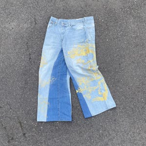 Op de afbeelding: Lichtblauwe en medium blauwe spijkerbroek met wijde pijpen en een contrasterend paneelontwerp. De voorkant van de broek is versierd met een grote, gedetailleerde gele grafische print van een draak die over de broekspijpen kronkelt.