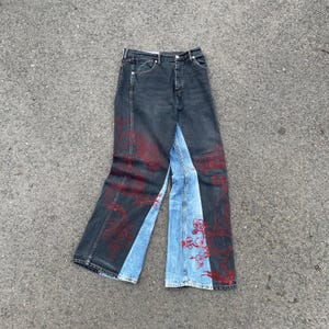 Puede incluir: Pantalones vaqueros de talle alto con un diseño de bloques de color en paneles de mezclilla gris carbón oscuro y azul claro. Las secciones gris oscuro están decoradas con intrincados dibujos de líneas rojas que representan ramas florales.
