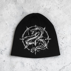 Peut inclure: Un bonnet en tricot noir avec une impression graphique blanche centrée sur le devant. Le motif représente un serpent enroulé à l'intérieur d'un cadre circulaire doté d'extensions pointues rayonnant vers l'extérieur, rappelant une étoile.