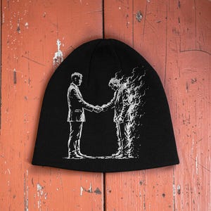 May include: A black knit beanie featuring a white graphic print of two figures in suits shaking hands. One figure is depicted in solid lines, while the other figure appears to be dissolving into flames or smoke, creating a high-contrast visual effect.