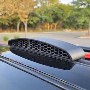 May include: A matte black, low-profile automotive roof vent featuring a distinct honeycomb mesh pattern. The curved, aerodynamic component is mounted directly onto the glossy black surface of a vehicle roof, showing a textured finish.