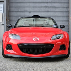 Può includere: Un'auto sportiva cabriolet rossa con tetto nero e dettagli neri. L'auto presenta un logo Mazda sul davanti ed è parcheggiata davanti a una porta di garage grigia.