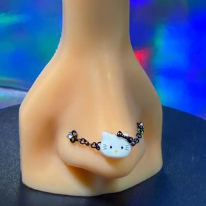 May include: A nose chain featuring a white Hello Kitty charm with a black bow and whiskers. The chain is black and adorned with small, clear gems. The jewelry is displayed on a mannequin nose.