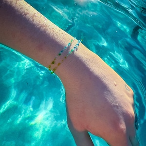 May include: A delicate bracelet featuring small, translucent beads in shades of blue, green, and yellow. The beads are strung on a thin, flexible cord, creating a subtle, layered appearance around the wrist with a light, airy aesthetic.