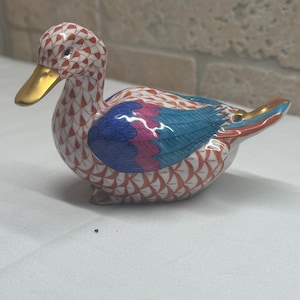 May include: A ceramic duck figurine with a gold beak and accents. The duck's body is white with a red and white pattern, and its wings are blue and pink. The figurine is a decorative item.
