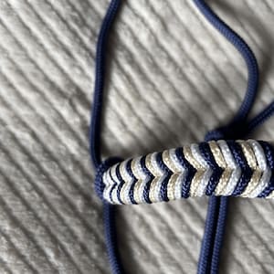 Peut inclure: Un licol de cheval bleu marine avec une muserolle décorative. La muserolle présente un motif en chevrons blanc et beige, ajoutant une touche élégante à l'équipement équestre. Le licol est fait de corde durable.