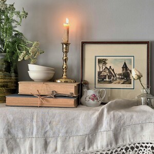 Peut inclure: Un bougeoir en laiton avec une bougie allum&eacute;e, deux livres beiges empil&eacute;s li&eacute;s par une ficelle, une cl&eacute; en m&eacute;tal, deux bols blancs, un petit pot &agrave; lait floral, une gravure encadr&eacute;e d'un village et une rose blanche dans une bouteille.