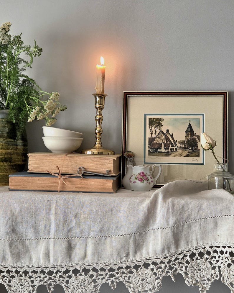 Peut inclure: Un bougeoir en laiton avec une bougie allum&eacute;e, deux livres beiges empil&eacute;s li&eacute;s par une ficelle, une cl&eacute; en m&eacute;tal, deux bols blancs, un petit pot &agrave; lait floral, une gravure encadr&eacute;e d'un village et une rose blanche dans une bouteille.