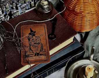 May include: A Queen of Roots tarot card featuring a figure in a pointed hat, placed on a stack of two books. Nearby are a small glass inkwell, a black metal key, a woven orange pumpkin-shaped basket, and a metal candle holder with a wax candle.