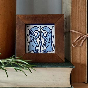May include: A square ceramic tile featuring a blue and white illustration of two stylized animals facing each other with a central floral motif, encased in a dark brown wooden frame with a smooth finish and mitered corners.
