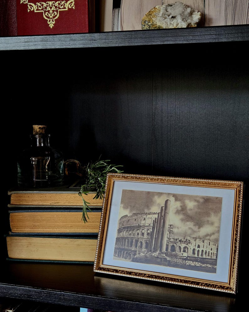 May include: A rectangular gold-toned picture frame containing a sepia-toned print of the Roman Colosseum. The frame features a textured, beaded edge and a white mat border. The artwork depicts the ancient stone amphitheater with prominent columns.
