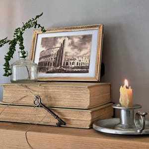 May include: A framed black and white print of the Colosseum sits atop two stacked beige hardcover books. A clear glass bottle with a green sprig, a metal skeleton key tied with twine, and a lit candle in a silver holder complete the display.