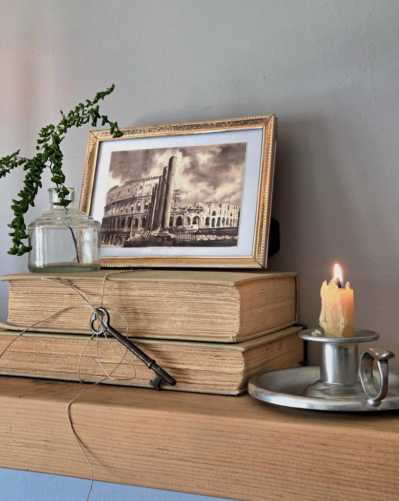 May include: A framed black and white print of the Colosseum sits atop two stacked beige hardcover books. A clear glass bottle with a green sprig, a metal skeleton key tied with twine, and a lit candle in a silver holder complete the display.