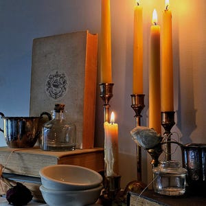 Peut inclure: Une collection de quatre bougies effil&eacute;es jaunes allum&eacute;es dans des bougeoirs en laiton, accompagn&eacute;es de deux livres anciens empil&eacute;s, une bouteille en verre transparent avec un bouchon en li&egrave;ge, deux bols en c&eacute;ramique blanche et un petit bocal en verre.
