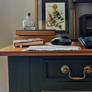 May include: A dark-painted wooden desk with a brass drawer pull, featuring a clear glass bottle with a cork stopper, two stacked books, a small patterned notebook with a pink ribbon marker, a black computer mouse, and a framed bird illustration.