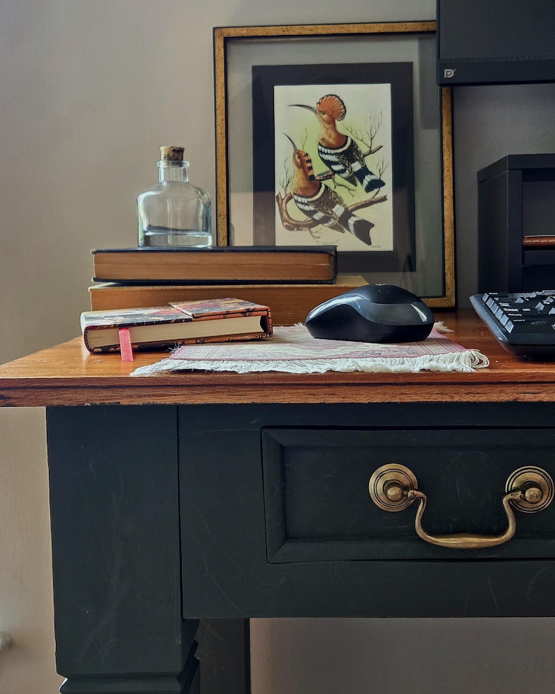 May include: A dark-painted wooden desk with a brass drawer pull, featuring a clear glass bottle with a cork stopper, two stacked books, a small patterned notebook with a pink ribbon marker, a black computer mouse, and a framed bird illustration.
