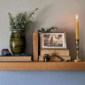 May include: A green ceramic vase with greenery, a purple amethyst crystal, two stacked books, a framed black and white print of the Colosseum, a small glass inkwell, a metal key tied with twine, and a yellow taper candle in a brass holder.
