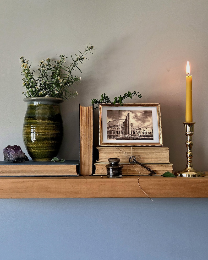 May include: A green ceramic vase with greenery, a purple amethyst crystal, two stacked books, a framed black and white print of the Colosseum, a small glass inkwell, a metal key tied with twine, and a yellow taper candle in a brass holder.