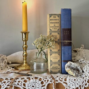 May include: A brass candlestick with a lit yellow candle, a clear glass bottle containing small white flowers, two hardcover books titled The Bounty Trilogy and The Spy, a white lace doily, and a small geode crystal on a wooden shelf.
