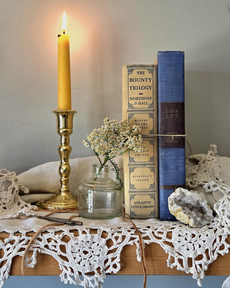 May include: A brass candlestick with a lit yellow candle, a clear glass bottle containing small white flowers, two hardcover books titled The Bounty Trilogy and The Spy, a white lace doily, and a small geode crystal on a wooden shelf.