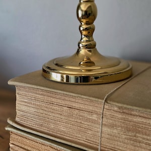 May include: A polished gold-toned metal candlestick base with a circular pedestal and a tiered, rounded stem, resting on top of two stacked, thick, beige-colored hardcover books with visible paper edges and a thin string wrapped around them.